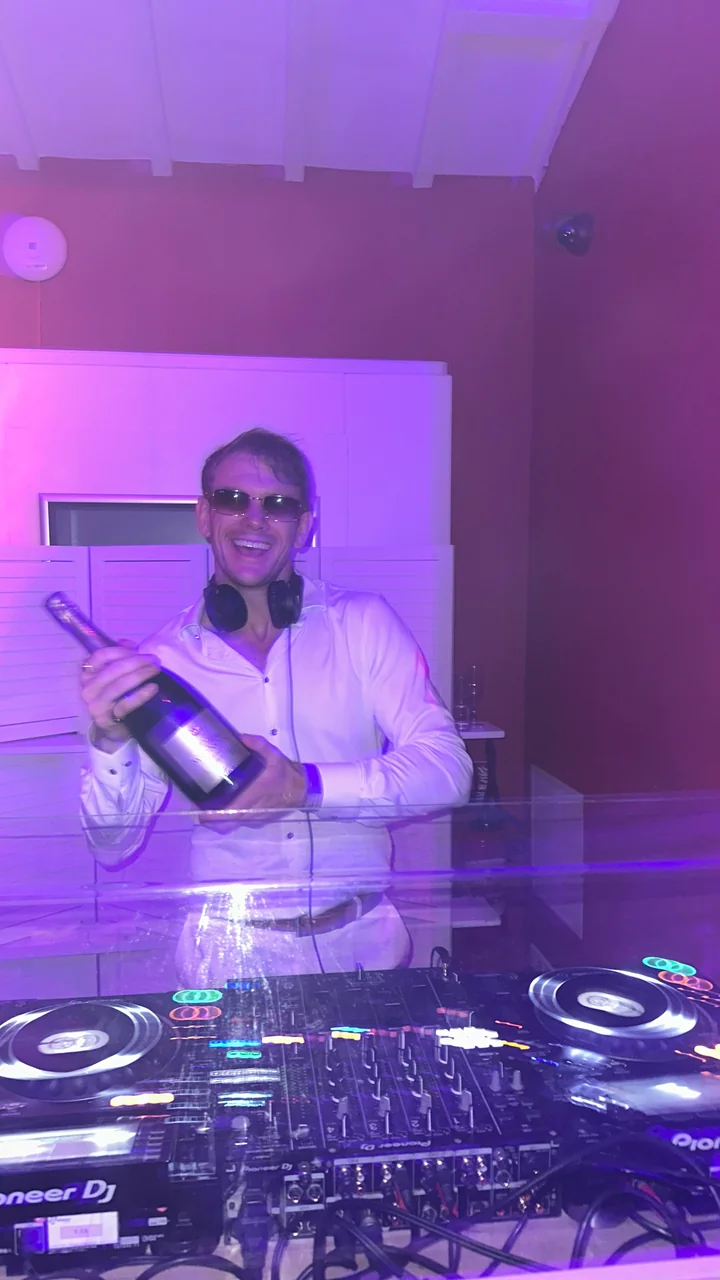 Joe smiling behind the DJ decks in a white shirt, with headphones around his neck.